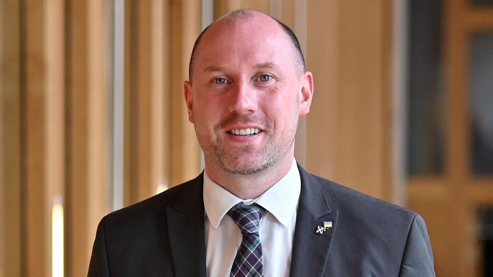 Head and shoulders shot of Neil Gray, who is smiling and has a bald head. He is in the Scottish Parliament building and is wearing a grey suit, white shirt and green, white and purple tartan tie.