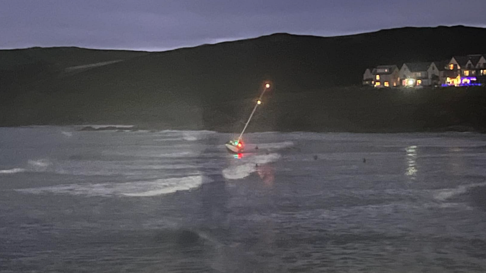 A small yacht with lights on the mast and hull lies at an angle on a beach as it gets dark with sea and beach visible below a row of houses with lights on.