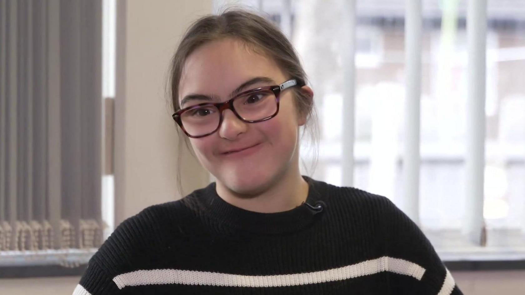 Laura has dark hair and dark rimmed glasses and is wearing a black and white striped jumper