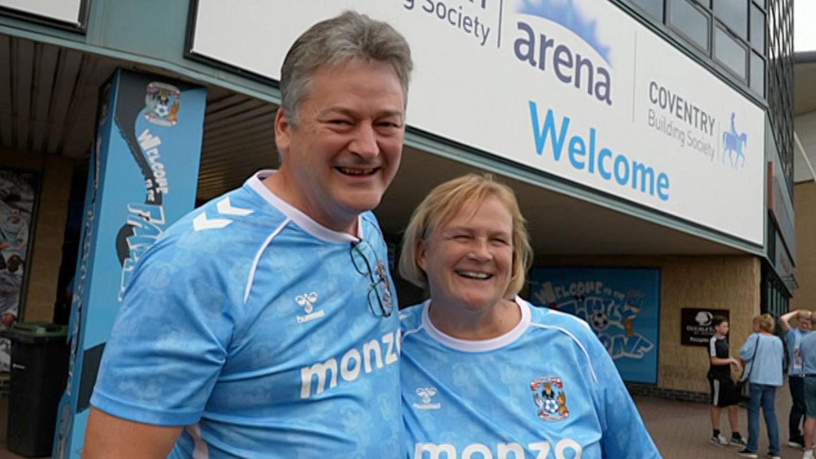A man and a woman are wearing blue football t-shirts that say 'monzo'. They are standing in front of the Coventry Building Society Arena.