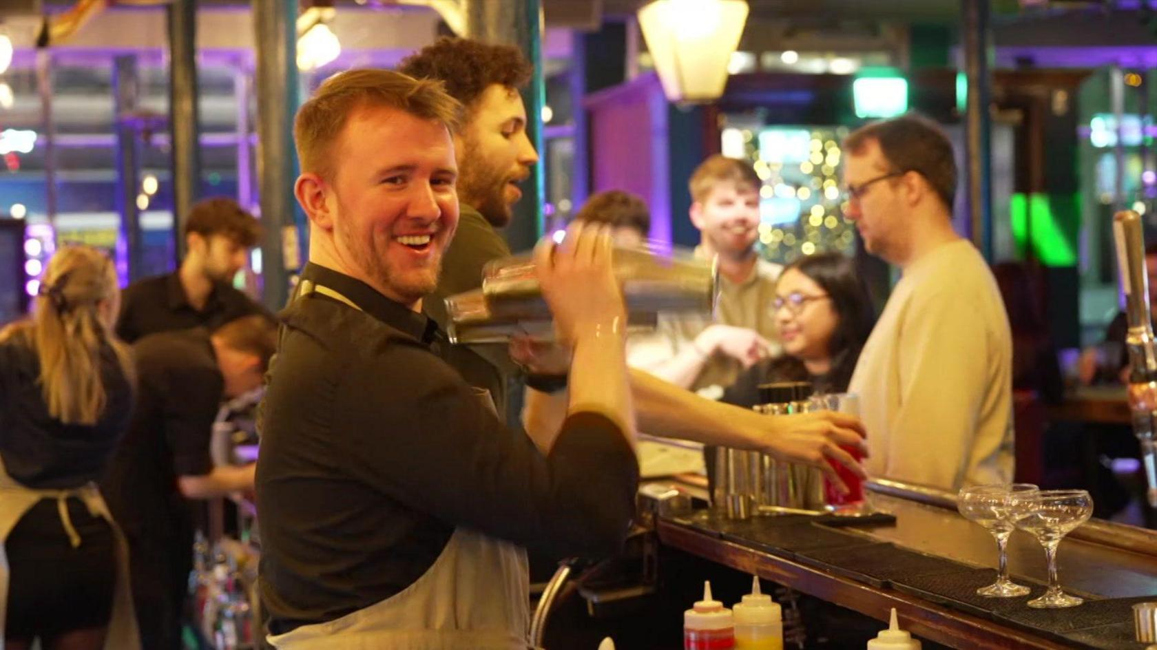 A male bartender stands behind a bar and smiles towards the camera while shaking a silver cocktail shaker in a brightly lit bar. He has short fair hair and is wearing a black shirt and beige apron. A colleague with short curly hair and a beard stands behind him serving a customer. Other customers and staff members can be seen in the background, slightly out of focus.