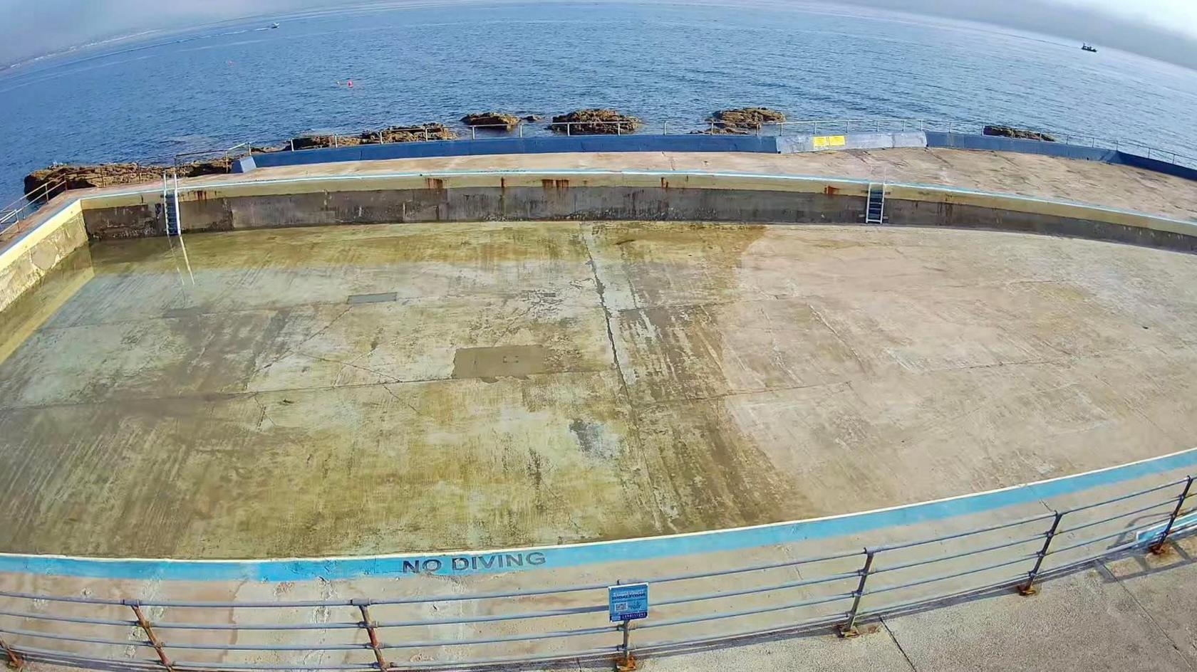 An empty concrete pool with a 'NO DIVING' warning painted near the bottom edge. A metal railing surrounds the pool, and a ladder is visible on the far side. In the background, the ocean stretches out with several rocks protruding from the water.