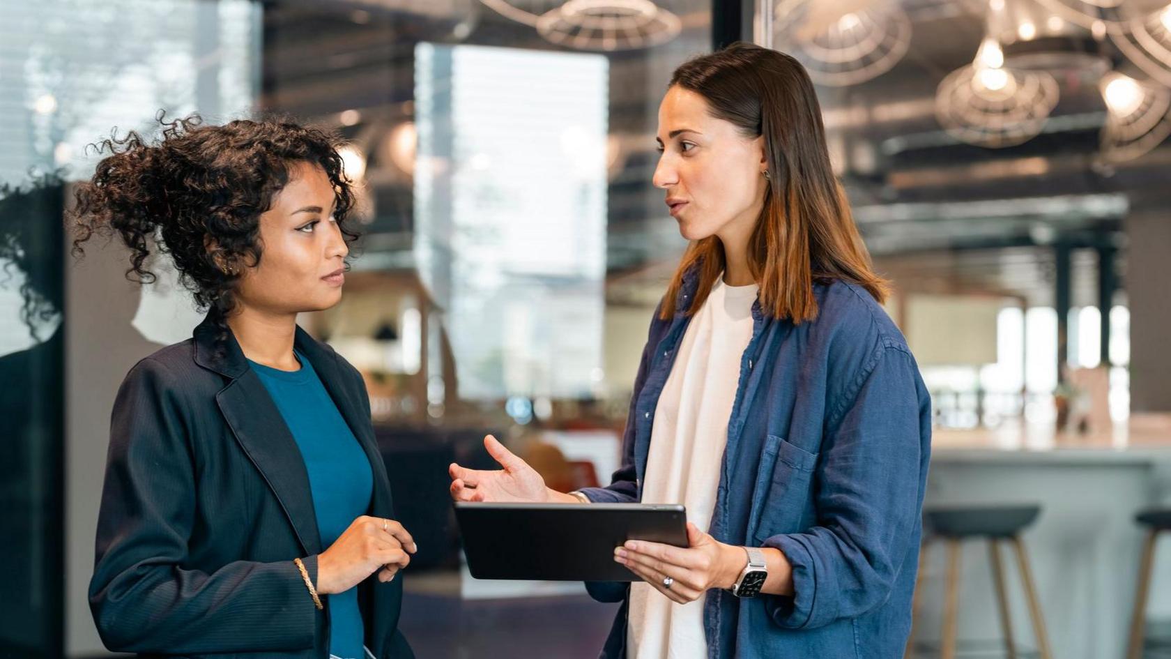 Two young business women working together, standing at modern office space room looking at a tablet computer. Both dressed in smart-casual business clothing.