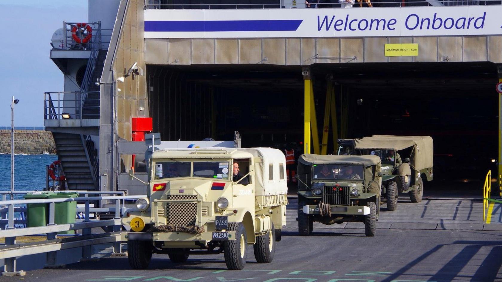 Three military vehicles, including a troop carrier and Jeeps, driving off a ferry ship.