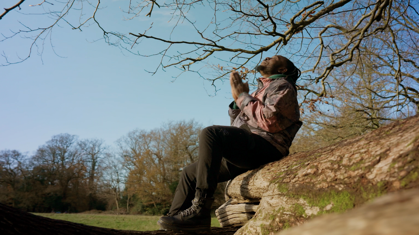 A man sat on a tree trunk. He is wearing a burgundy and purple coat and black jeans. He is sat with his hands together and is looking up towards the sky 