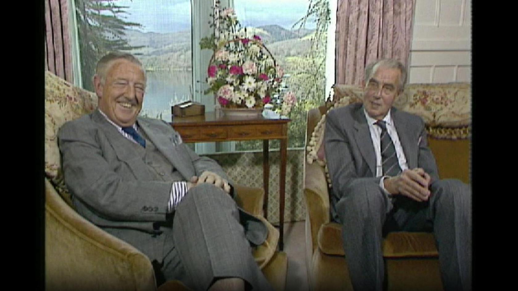 A screenshot from an archive interview filmed at Sharrow Bay. Brian Sack and Francis Coulson are both wearing grey suits and are sat on velvet armchairs inside the hotel. Behind them is a window giving onto Ullswater, with a view across the Lake.