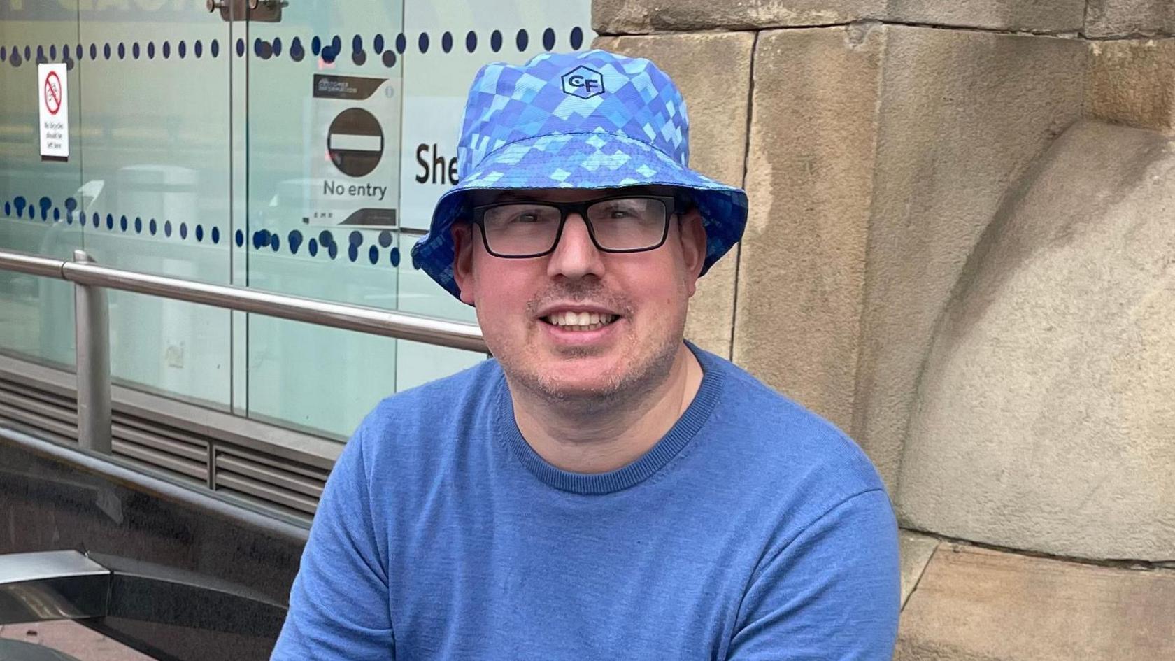 A man smiles at the camera. He wears a blue t-shirt and a bucket hat in a similar shade, patterned with squares. He has square glasses on and is sat on a public bench, just outside Sheffield railway station.