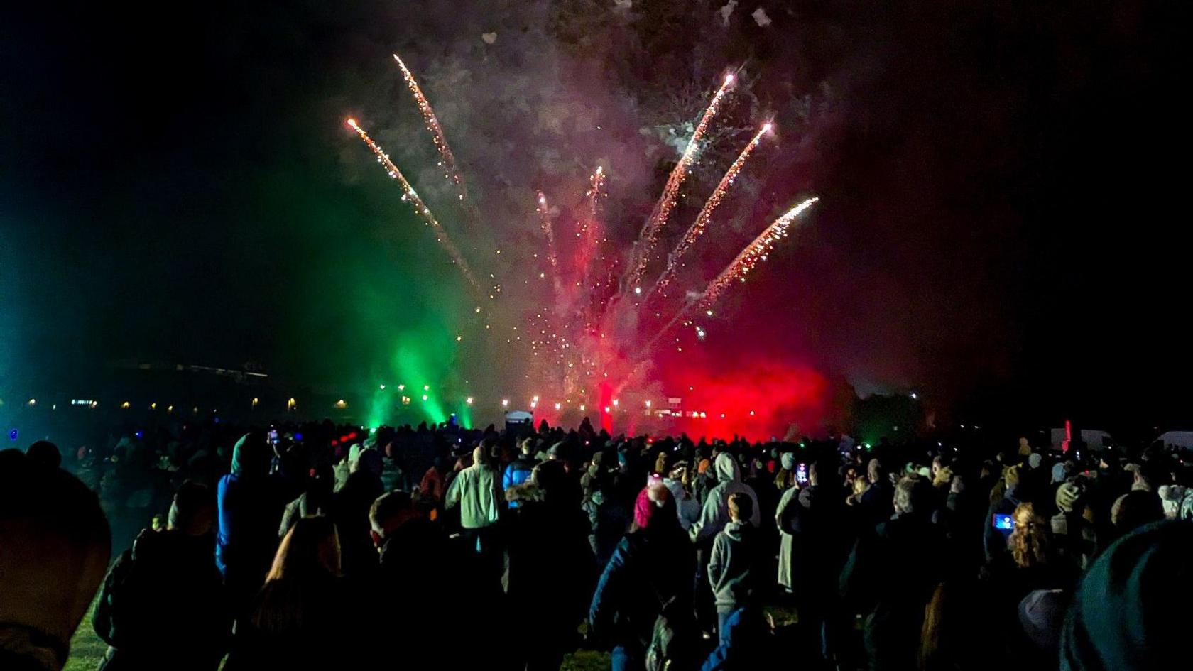 Red and green fireworks explode over a large crowd at night, at the Downend Round Table Fireworks Display near Bristol. Rockets are sending multiple spark showers into the sky