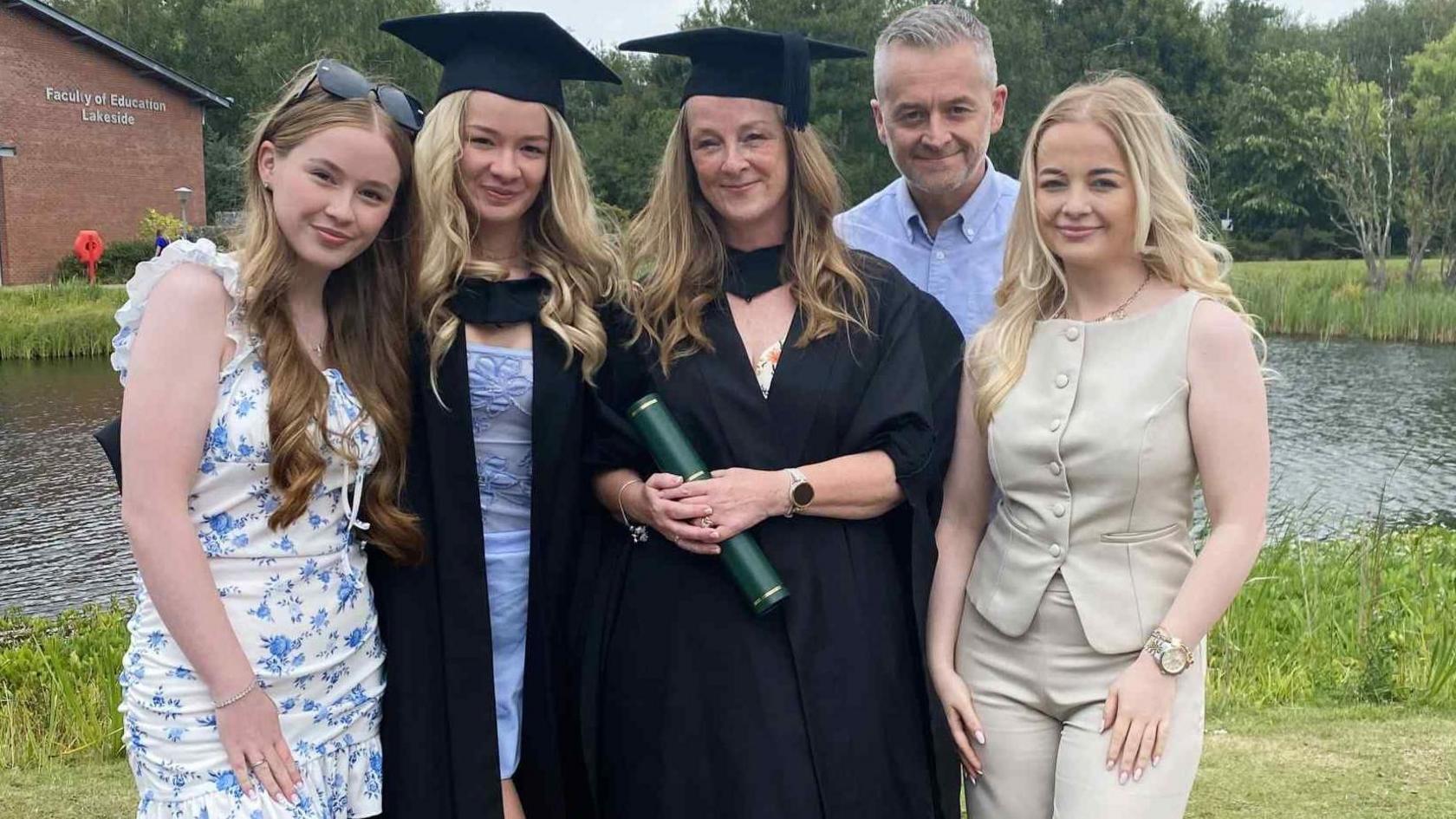 Stephanie Kennedy and daughter Mia both have shoulder length blonde hair and are wearing black mortar boards and academic gowns. He other two daughters also have long blonde hair - one is wearing  white dress with blue flowers and the  other wears fawn trousers and matching sleeveless top. Her husband in the background has greying and is wearing a sky blue Oxford shirt