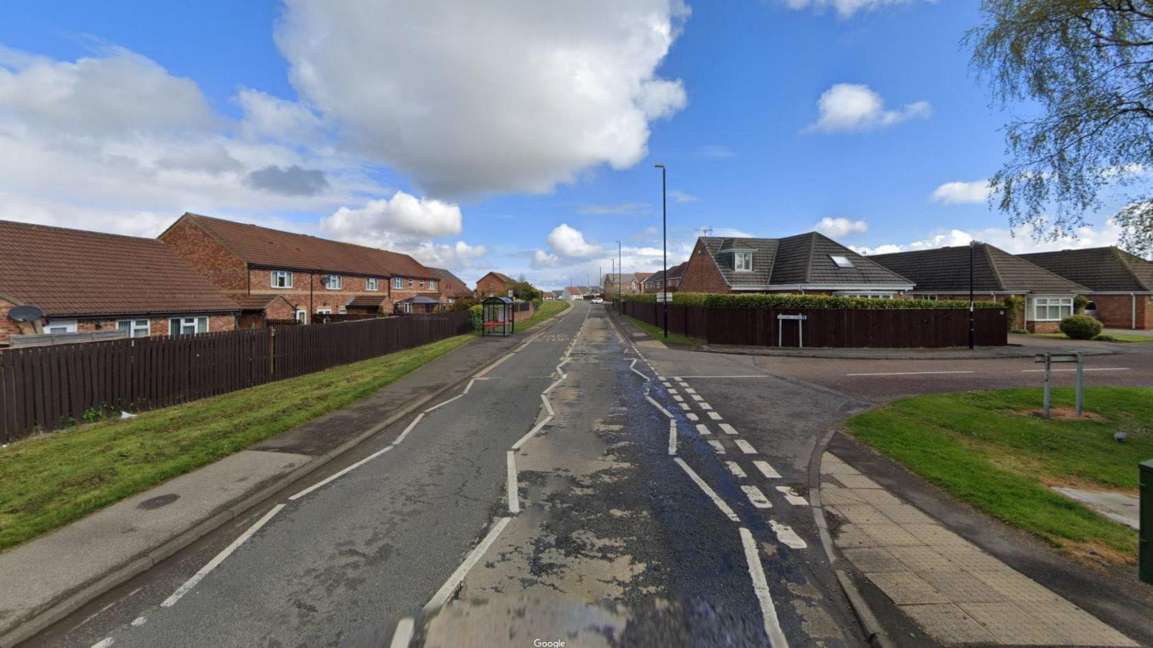 Burdon Road is a single-carriageway road with red-bricked houses behind brown fences bordering it. A metal bus stop is on the left-side pavement. The junction onto Lidcombe Close is on the right.