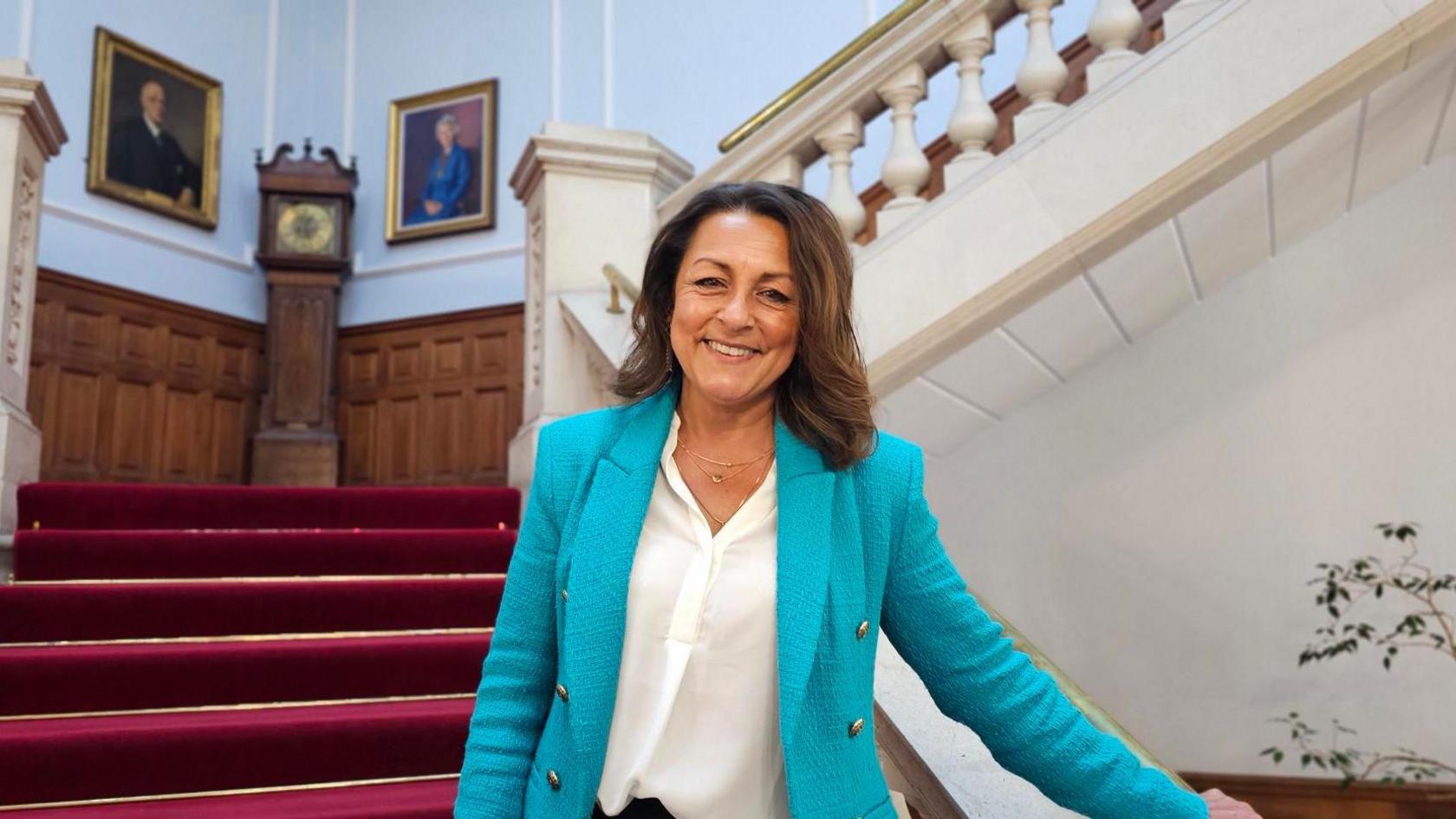 Linden Kemkaran stands on a staircase and holds the bannister with one hand. She is wearing a blue suit jacket and smiling at the camera. Behind her the staircase is covered in a red carpet and a grandfather clock stands on a landing area.