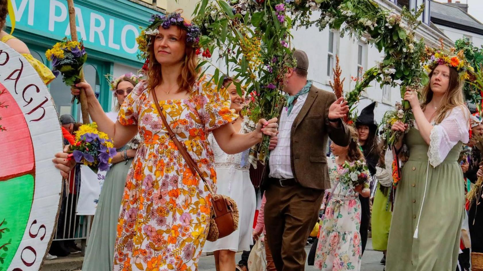 A woman in a orange and white floral dress leads a group of people carrying floral decorations forming a float in the parade.