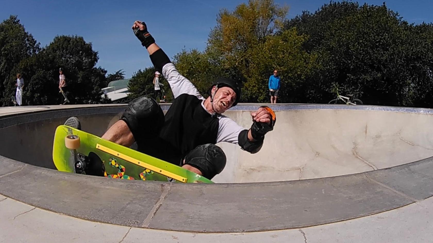 The picture shows a person skateboarding in a concrete skatepark bowl. They're equipped with full protective gear—helmet, knee pads, elbow pads, and wrist guards. The skateboard has a green deck with colourful graphics underneath. In the background, there are other people around the skatepark, along with trees and a clear blue sky.