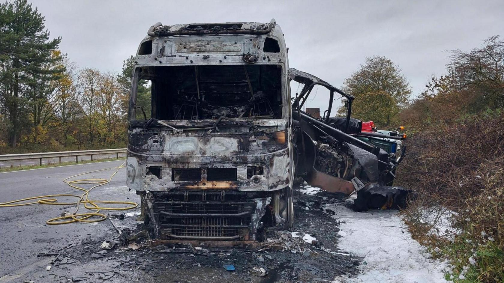 The front of a burnt out lorry following a fire. The road is severely damaged and debris surrounds the vehicle