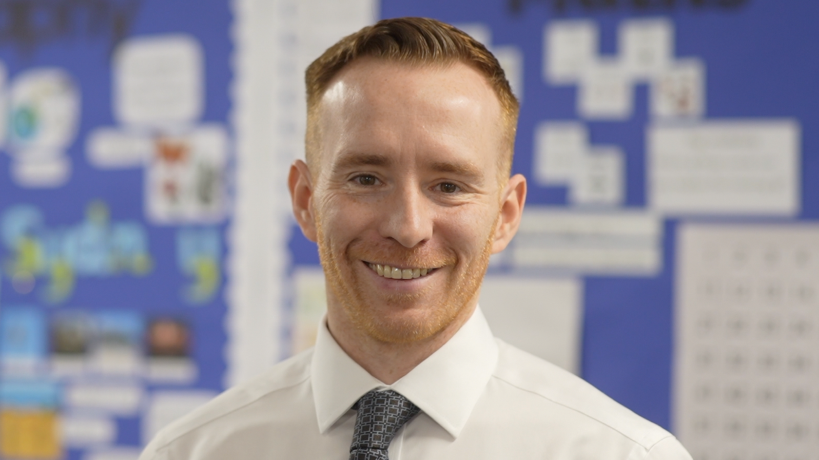 Head of school Julien Mealey in front of a blue board in a classroom which has work displayed on it. He has short ginger hair and stubble and is smiling while wearing a white shirt and dark tie.