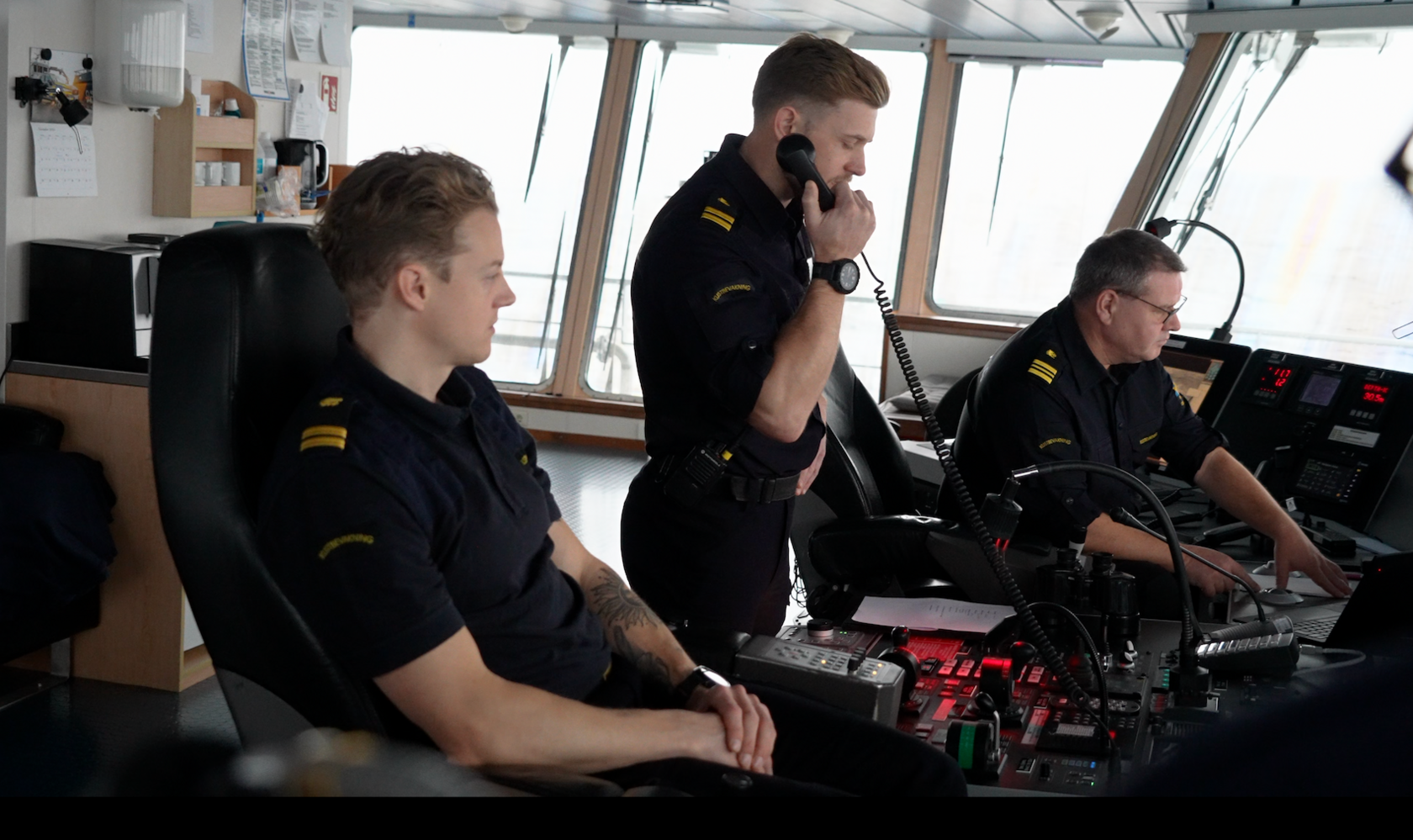 Three men in the Swedish coastguards uniform sit at the control desk of a ship. Two on the outside are sitting down while the man in the middle is standing and speaking on the phone.