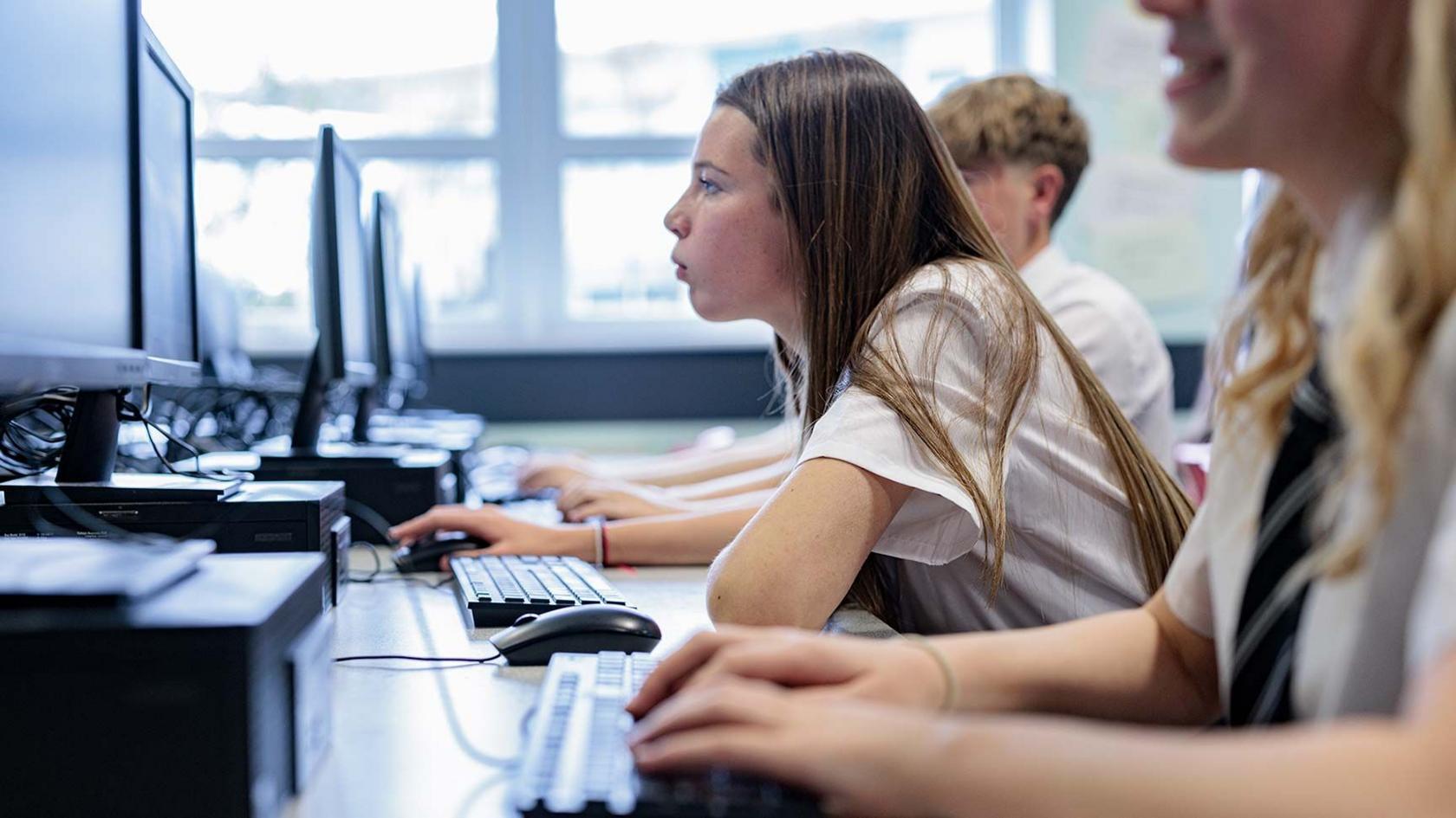 Profile of a teenage girl with long hair in school uniform in a classroom looking closely at a computer screen. Fellow students sit either side of her.