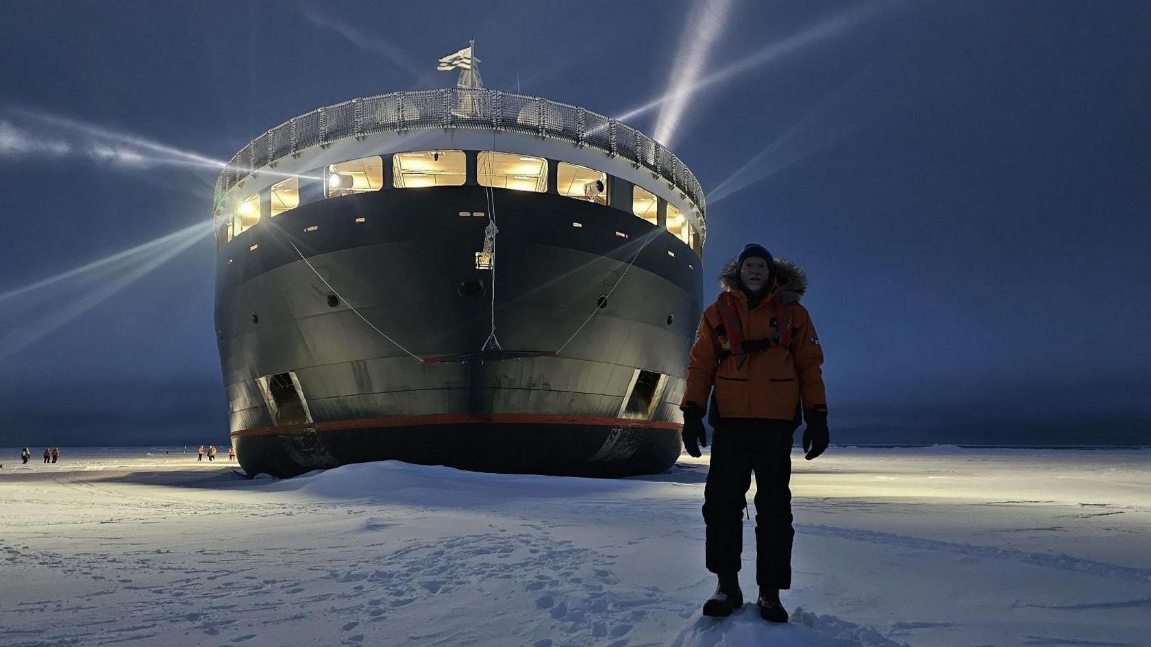 A man stands in the dark in front of a huge ship. He is wearing a big orange puffer coat, hat and gloves. There is ice all around.