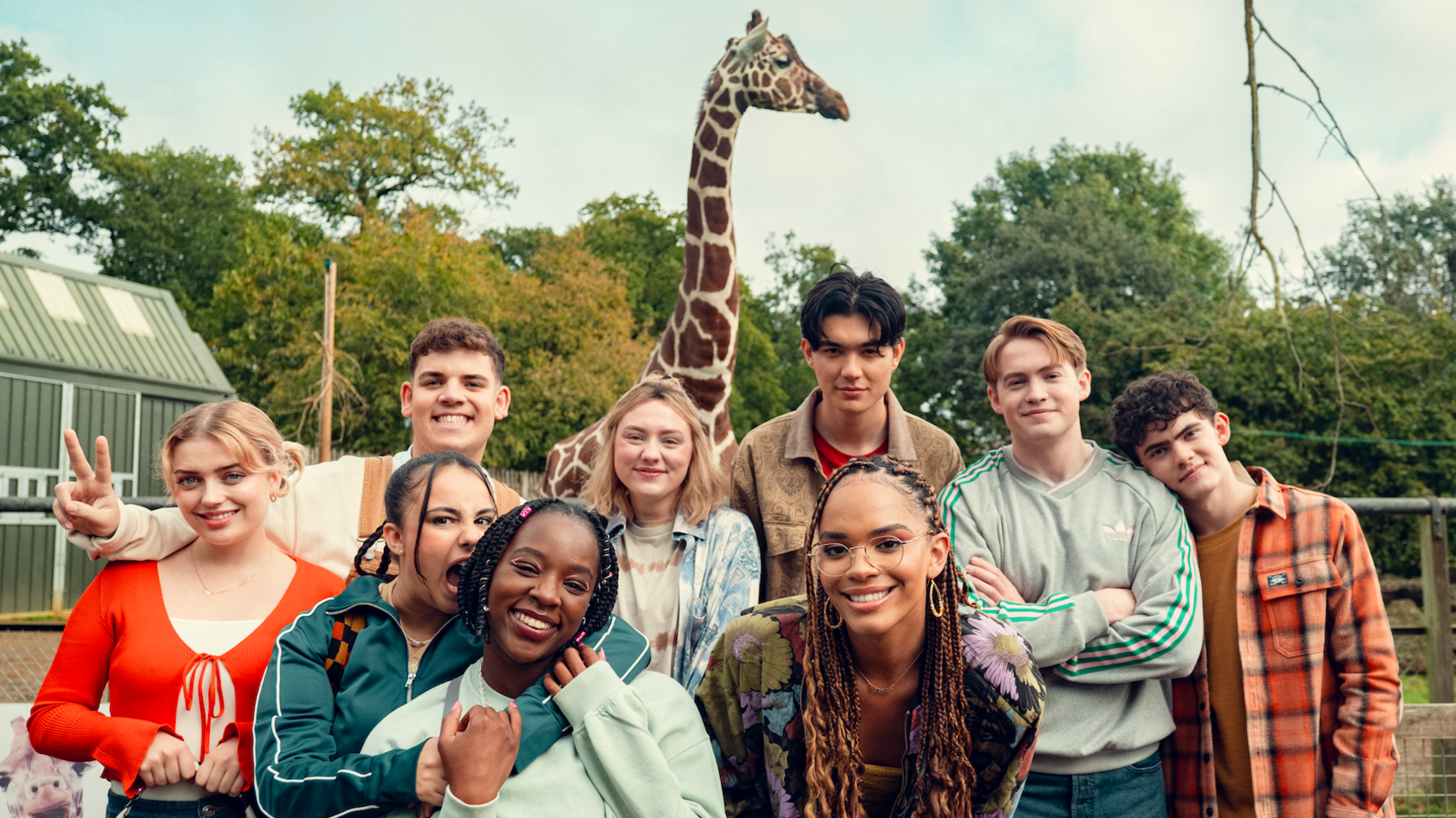 The cast of Hearstopper smile at the camera while filming on location at a zoo. A giraffe and trees are visible in the background.