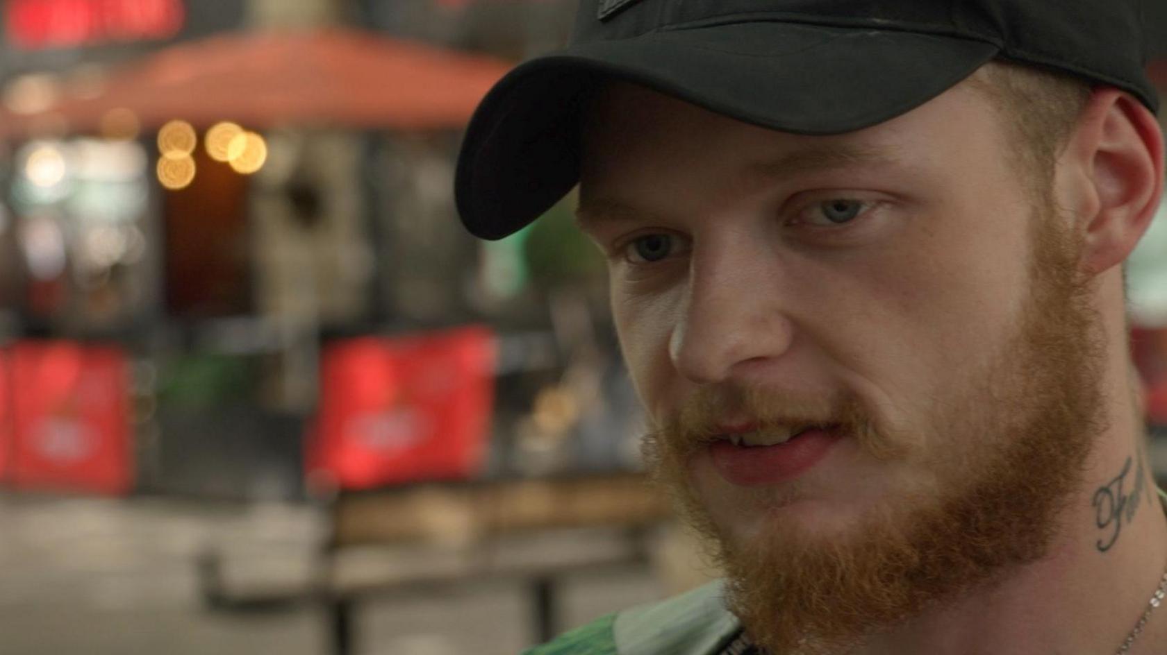 Lennie has a beard is wearing a black baseball cap. He is standing outside, with a street behind him.