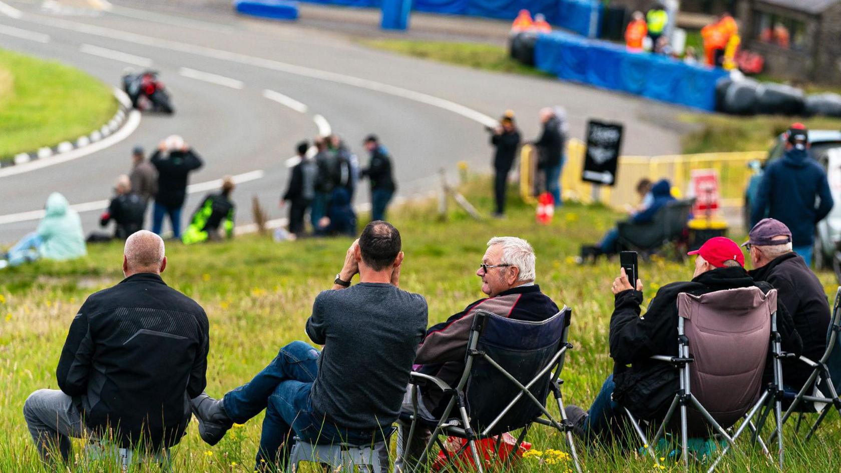 Spectators sitting in a row in camp chairs watch a motorcycle round a bend on the Mountain Road. They are on a bank of green grass and there are blue crash barriers at the side of the road in the distance.