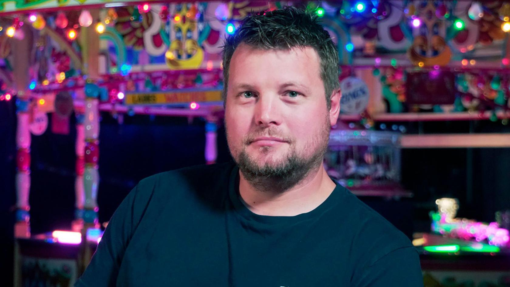 Raecher with short hair and short beard looks at camera in front of ferris wheel