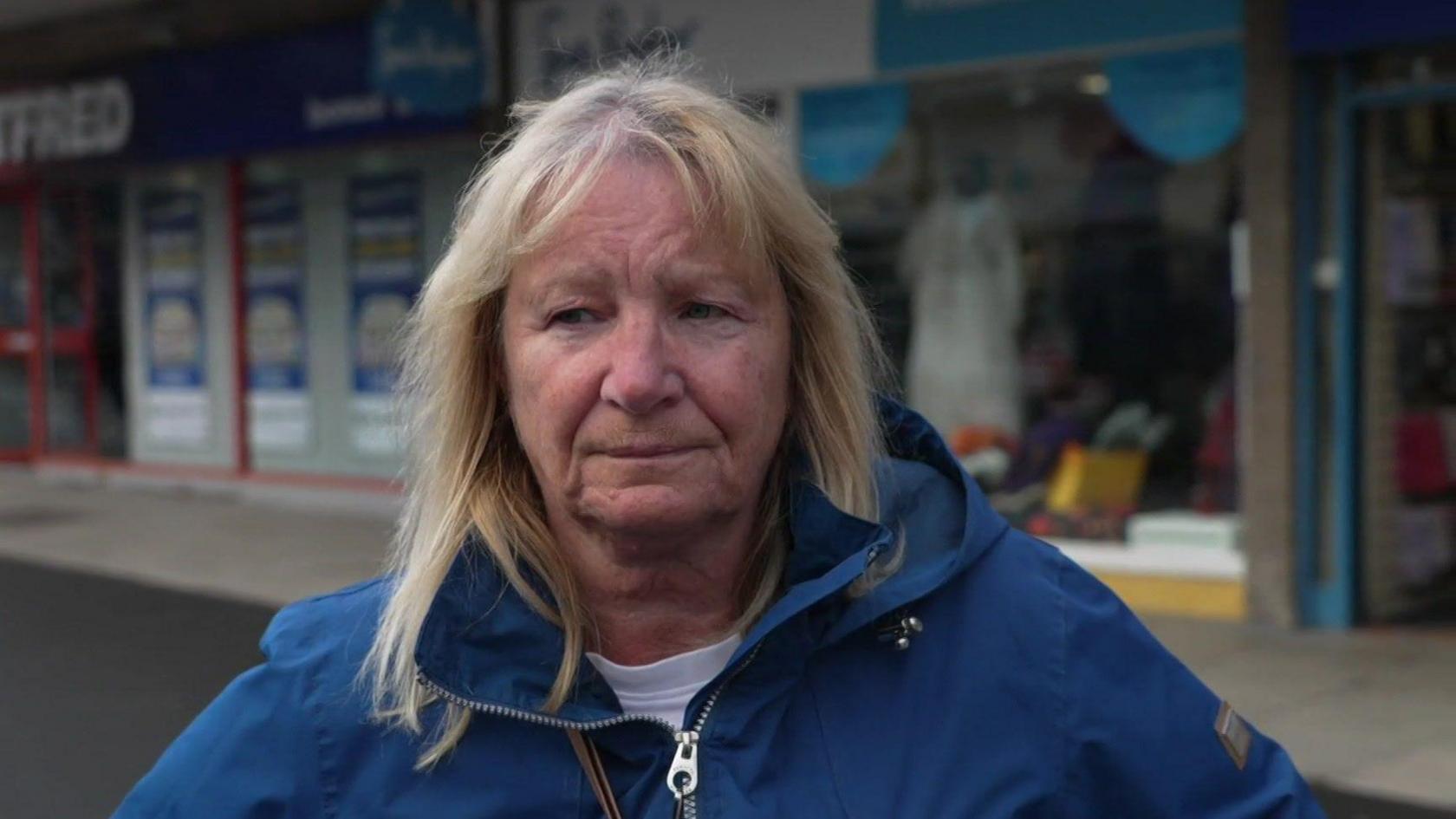 Image shows a lady with shoulder length blonde hair. She is wearing a blue jacket zipped up and a white top.  She is stood in front of a row of shops on Pudsey high street.