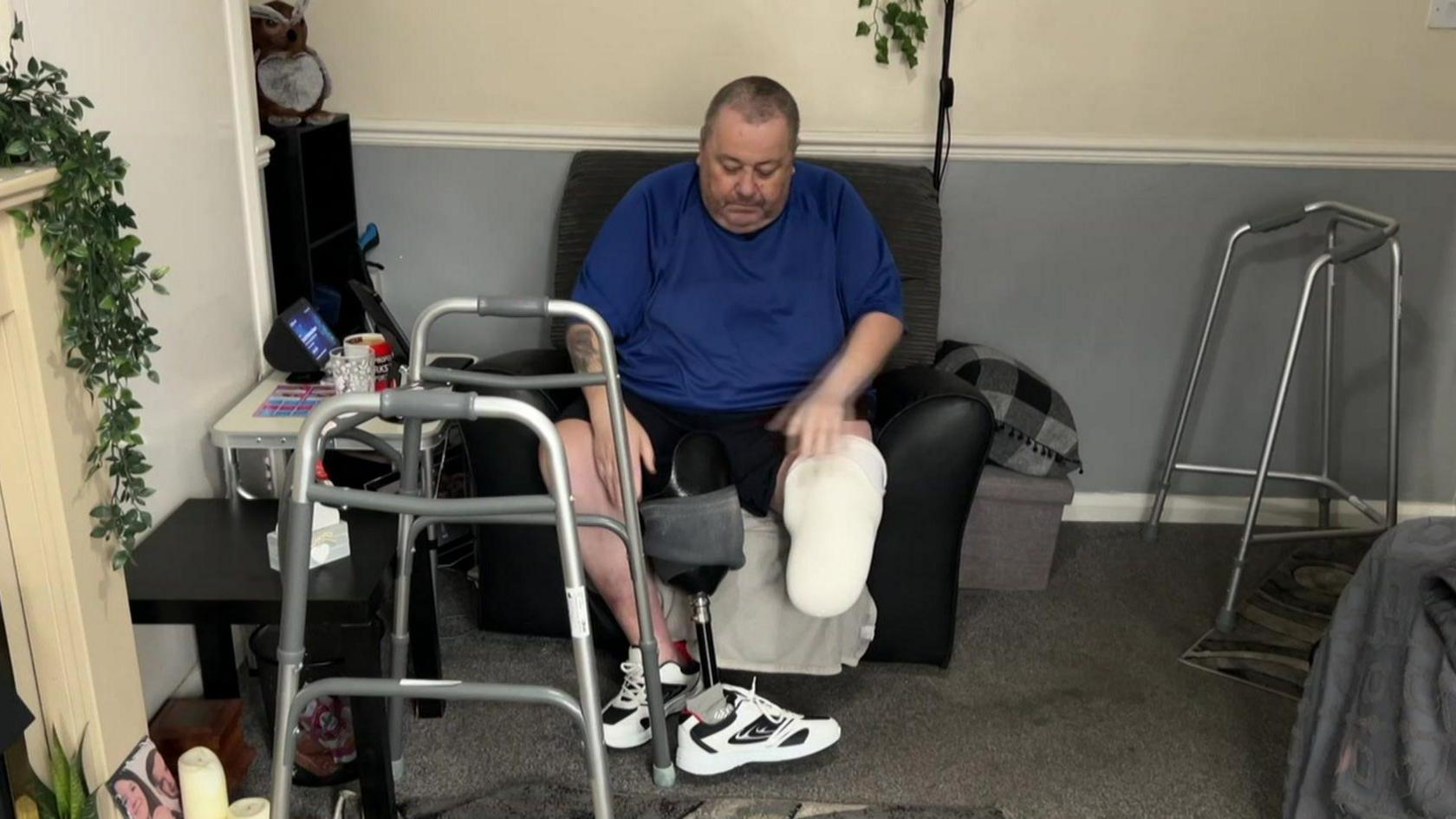 A man sits in a chair strapping a prosthetic to his left leg. There are grey carpets and black furniture in the room. He also has two walking aids.