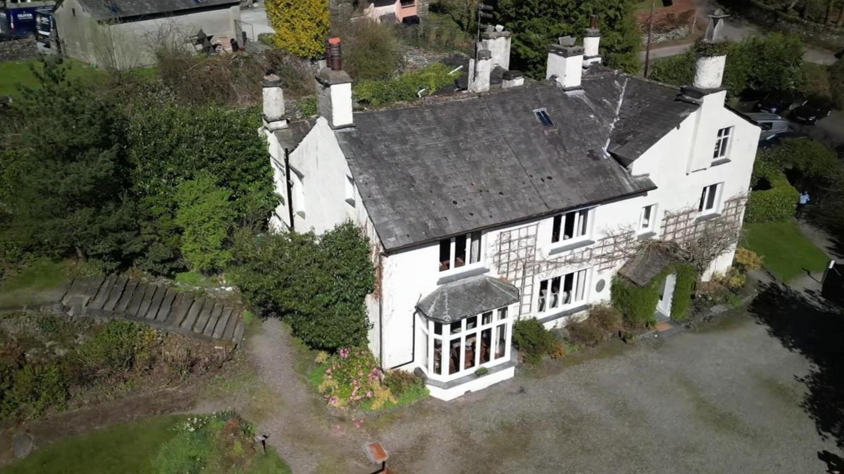 An aerial view of the large, white house on a sunny day. There are trees next to it as well as what looks like a farm.