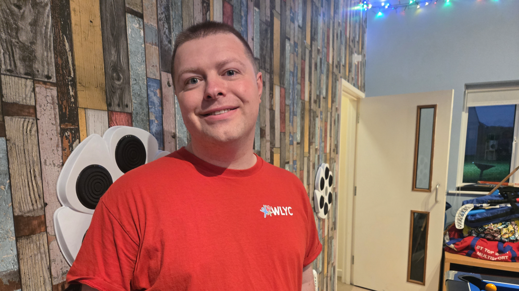 A man with short brown hair is wearing a red t-shirt branded with the name of the youth centre 'WLYC'. He is smiling at the camera. He is standing in front of a wooden wall and in the background is a desk with sports equipment piled on it.