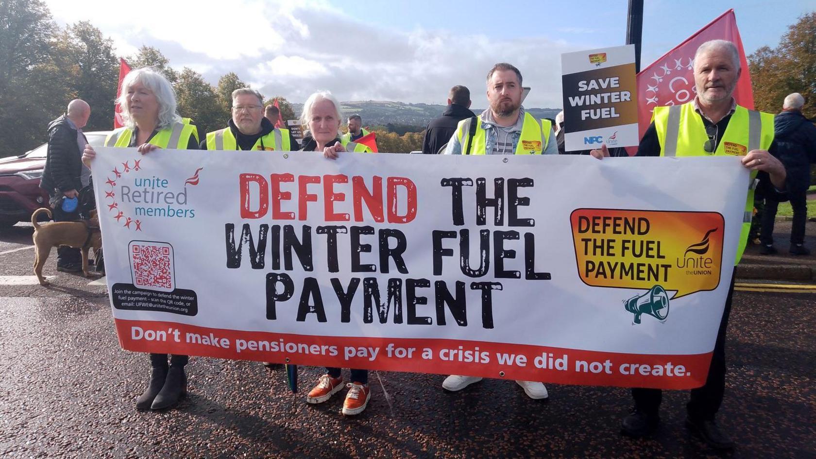 Five people wearing yellow high visibility jackets are holding a banner which says defend the winter fuel payment. Other placards can be seen in the background saying save winter fuel.