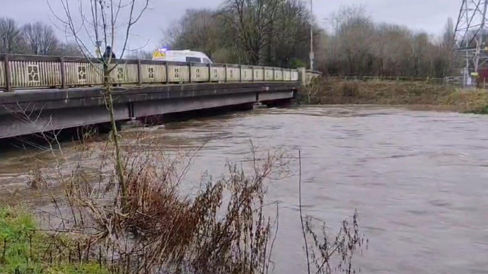 Very high flood water close to the bottom of a bridge crossing the river. A police van is parked on the bridge