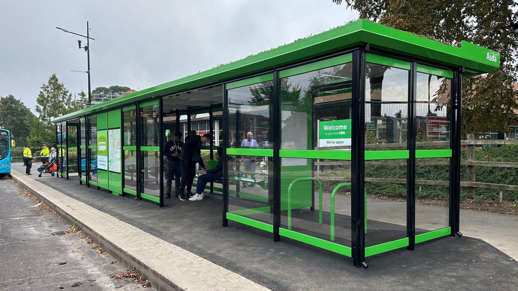 A bright green bus shelter on a pavement with a bus in the distance