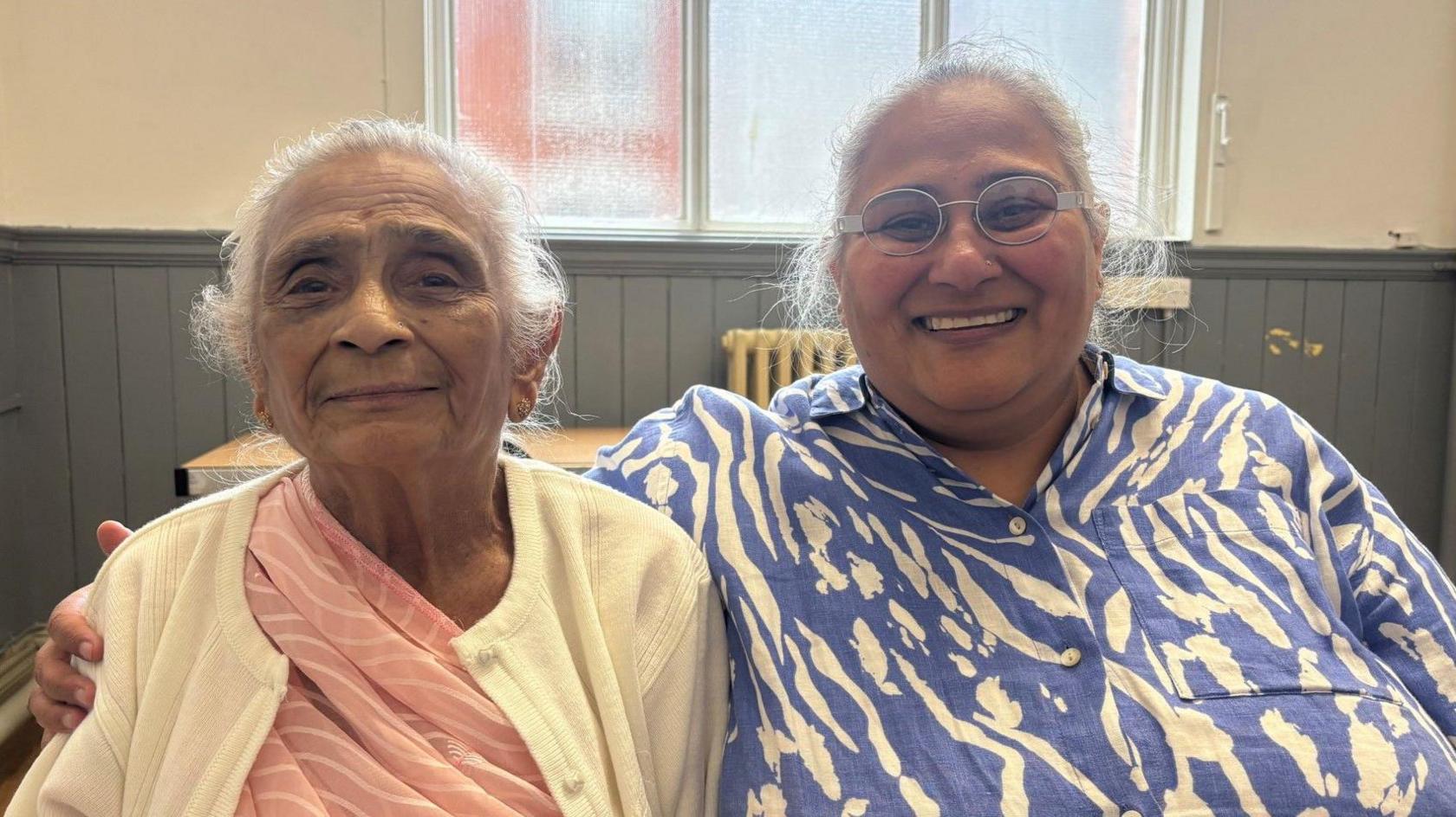Hindu mother and daughter smiling at the camera. Daughter is hugging her mother as they both sit and show the warm bond they share. Mother is wearing traditional attire, a pink a saree with a cream cardigan. The daughter is captured wearing a blue and white patterned dress as they sit in an empty room but looking cosy highlighting themes of family, love, and happiness.
