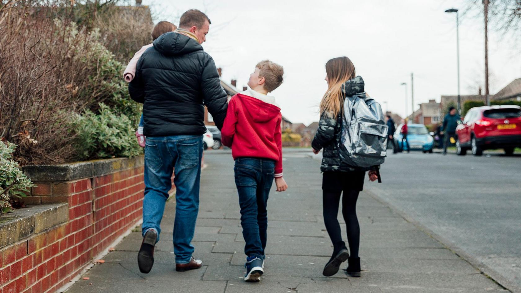 Father with 3 kids walking along the pavement, walking away from the camera. They are on a residential street in the UK 