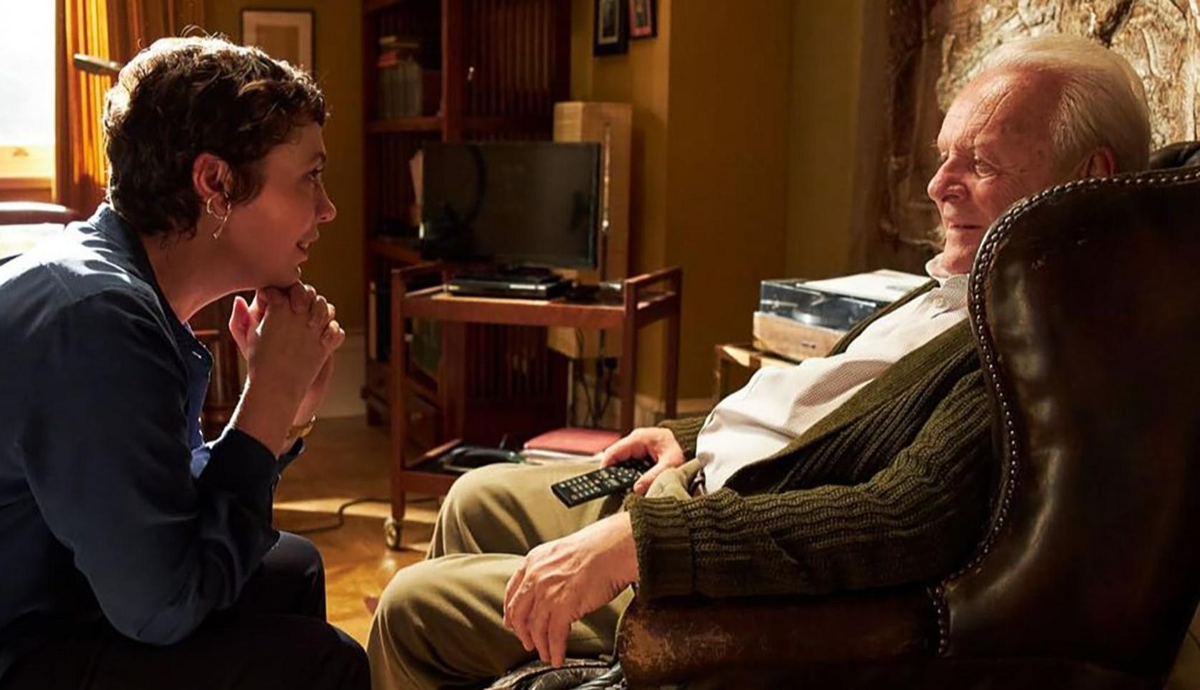 Olivia Colman closest to screen on left with her back to us. Profile. seated smiling with her hands on her chin. Short hair and gold looped earrings.
Hopkins smiling looking at her in arm chair. Also profile. Holding a tv remote.
They are in living room with tv set and record player in background