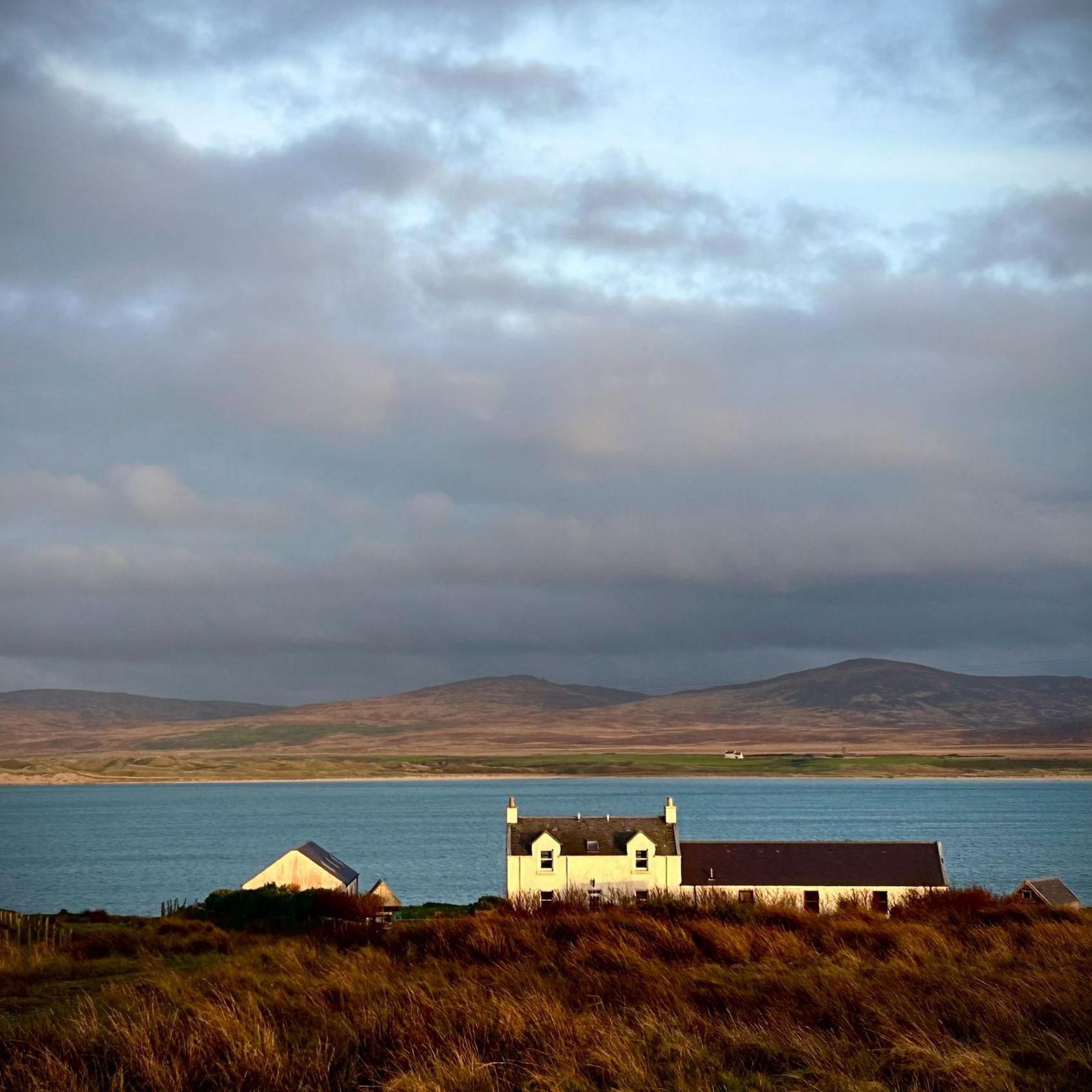 A white-walled two-storey house with a single storey extension attached sites on the shores of the loch. There are low hills in on the opposite shore.