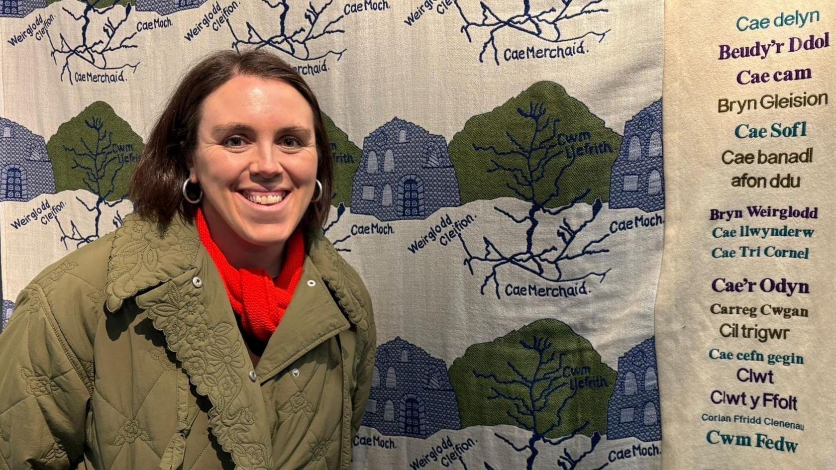 Naomi Jones yn sefyll o flaen carthen wlan gan wenu. Mae ganddi siaced frown a sgarff goch ymlaen. Mae ganddi wallt brown byr.