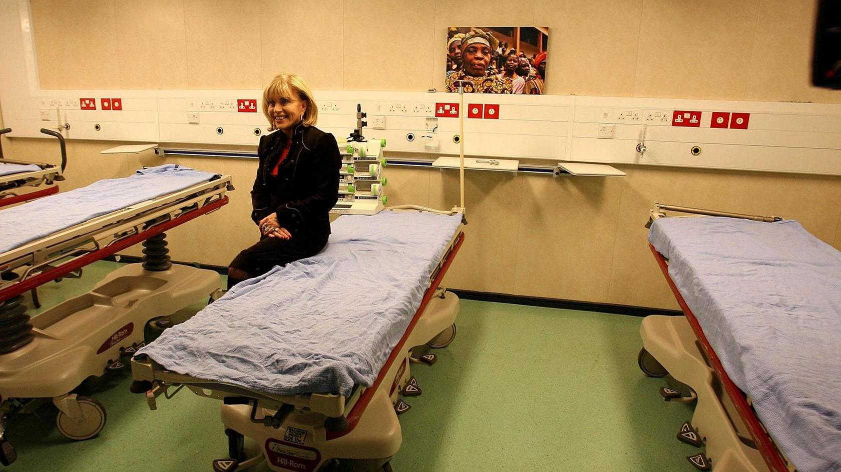 Dame Ann Gloag sitting on a bed on the Africa Mercy hospital ship, with several other beds nearby.