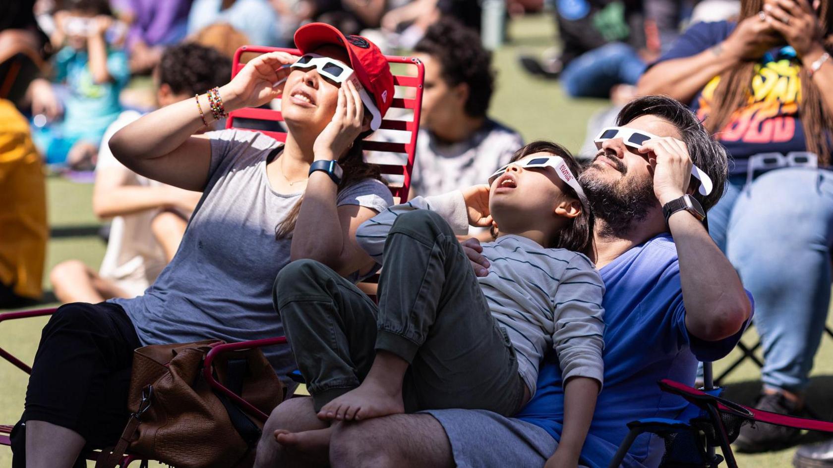 A man, woman and young child sit on deckchairs, wearing paper glasses with dark lenses. They look up to the sky, where a shadow falls over them. 
