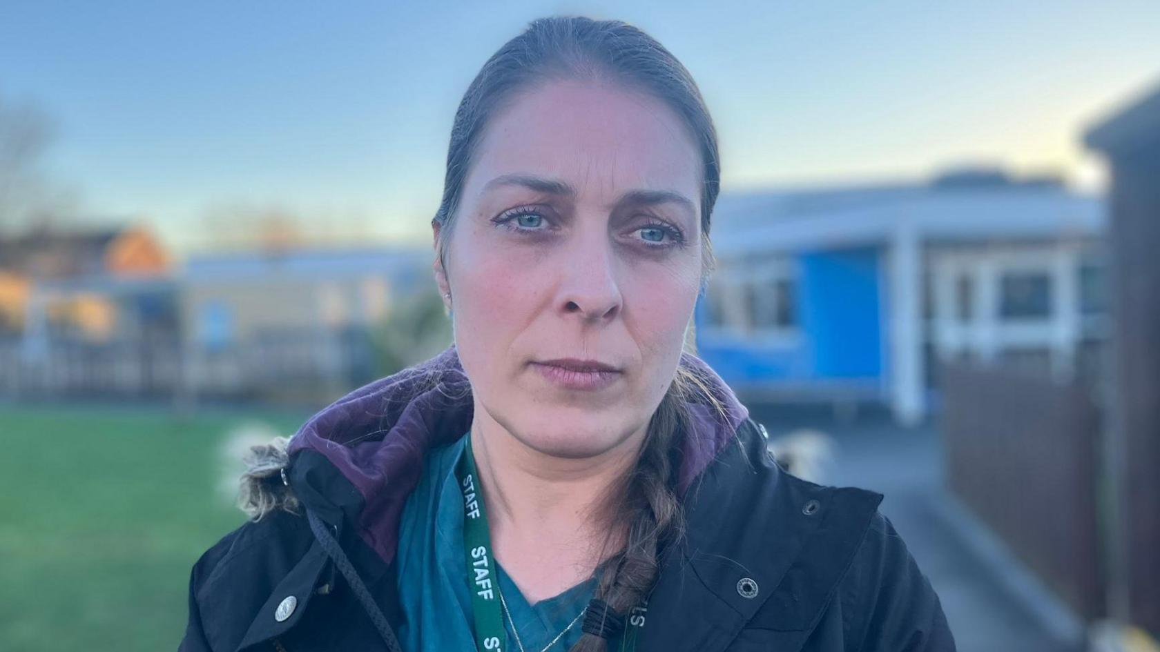 Mum, Sibel Ansell looks at the camera. She is wearing a green top and black coat. Her brown hair is in a side plait. The school is in the background.