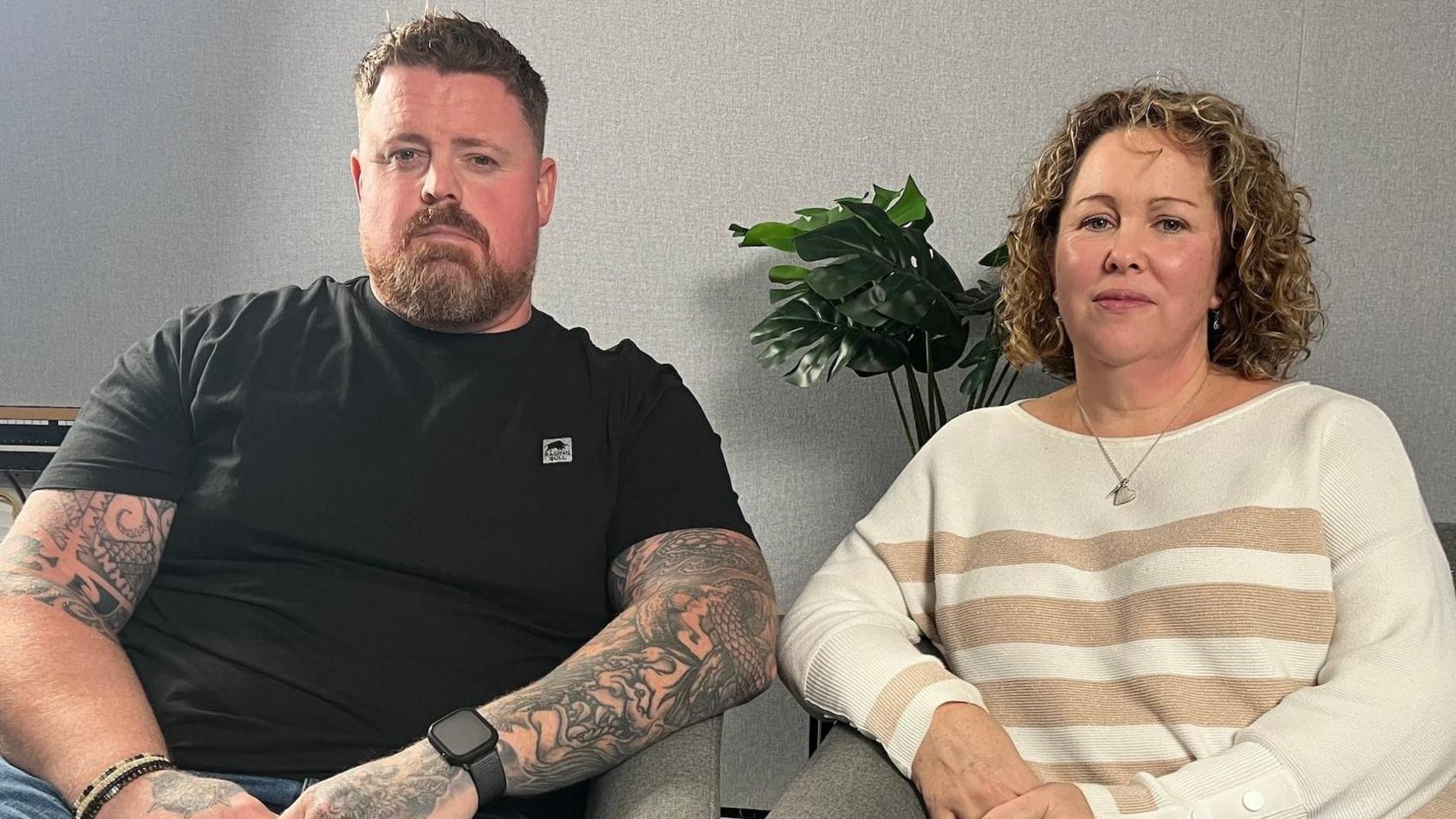A man with a black T-shirt and tattoos sits next to his wife who has curly hair and a beige jumper on. They are sitting in a grey studio. 