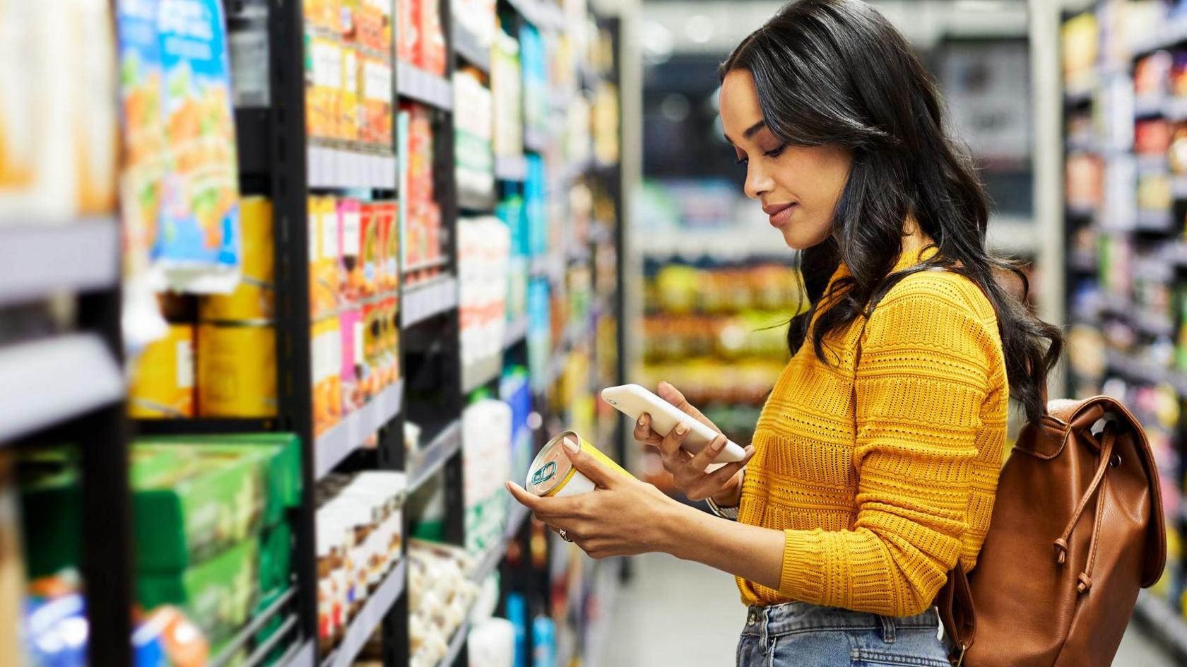 A woman wearing a yellow jumper looking at a tin of food while being on her phone in a supermarket 