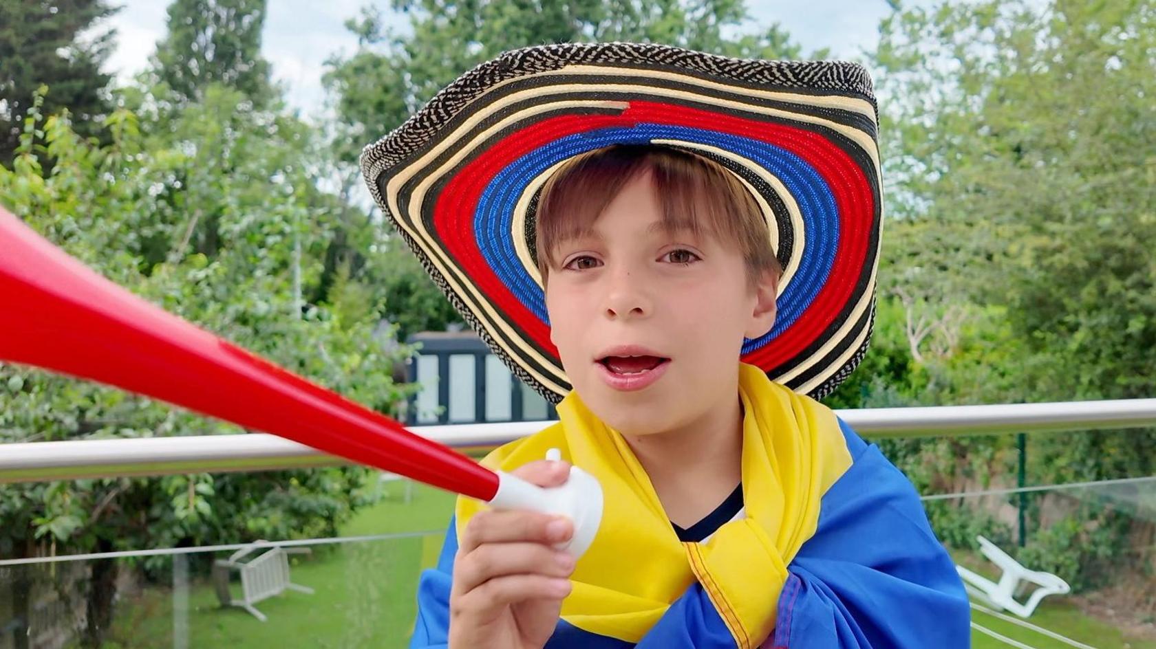 A boy wears a red, blue and black patterned hat and holds a red plastic horn.
