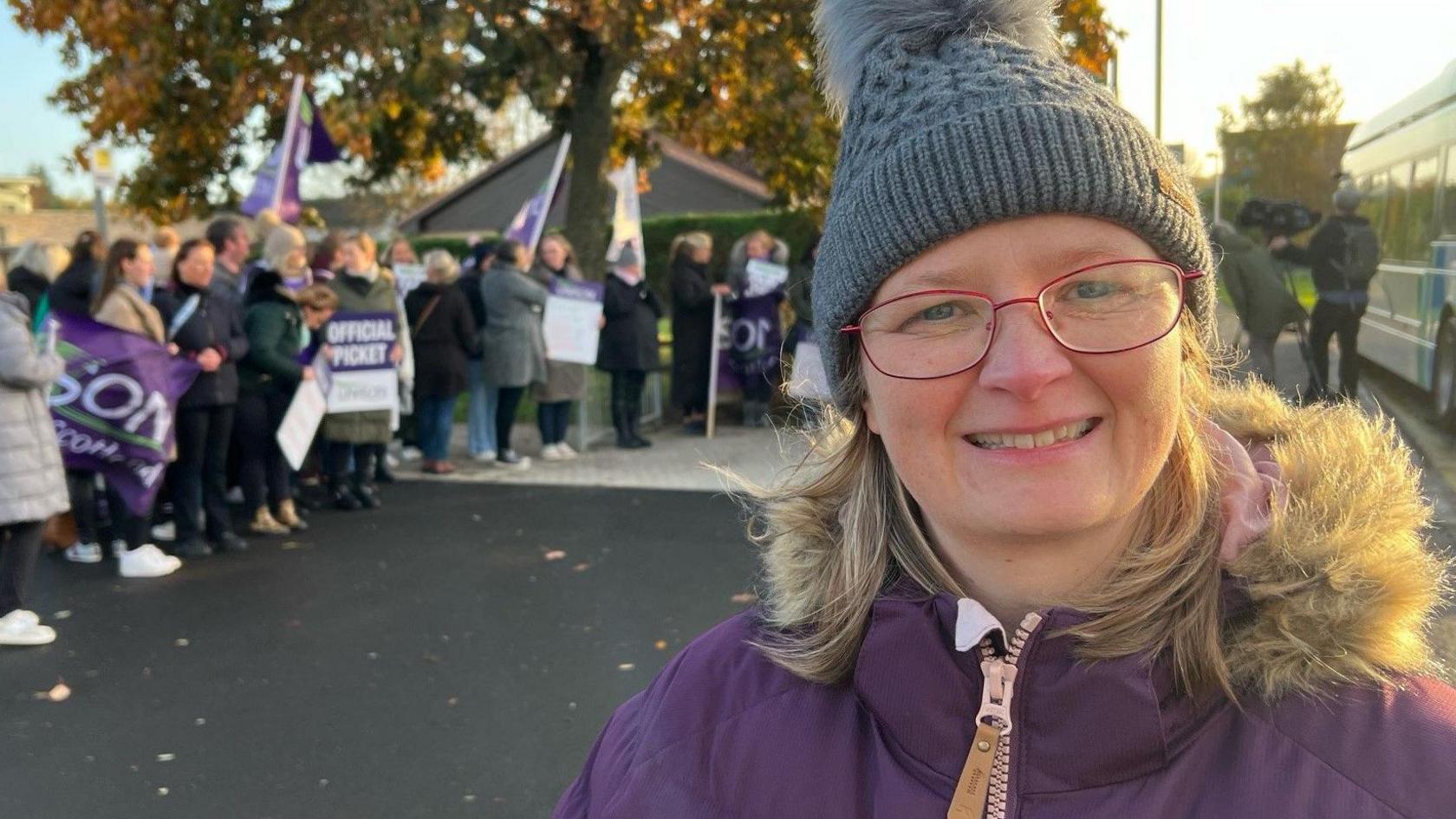 Terri Higgins wearing a grey bobbly hat and purple winter coat beside a picket line of striking workers