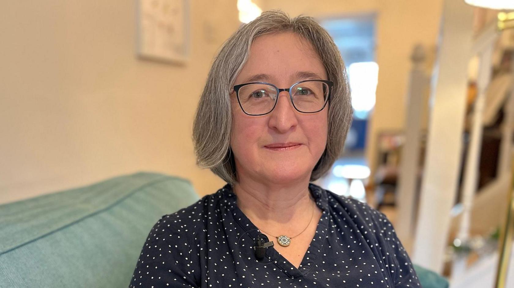 A woman with grey hair and glasses looks at the camera while sitting on her sofa. She is wearing a dark blue blouse with small white spots and is wearing a necklace with a pendant.