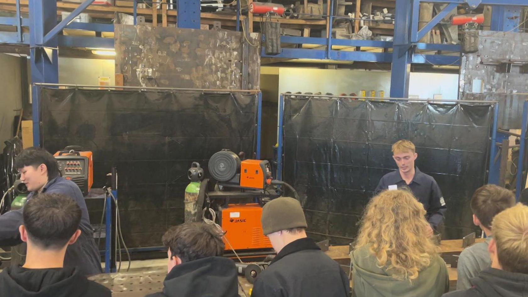 Two apprentices show a group of students some of the tasks they do, large machinery surrounds them.