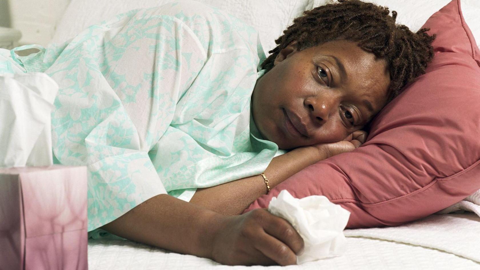 A woman lies in bed looking unwell holding a tissue with a box of them nearby