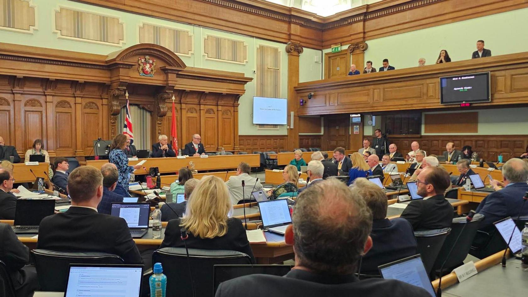 Members of Kent County Council sit in the debating chamber of County Hall, Maidstone, a large oak panelled room, with each member having a microphone and laptop in front of them on their desks.