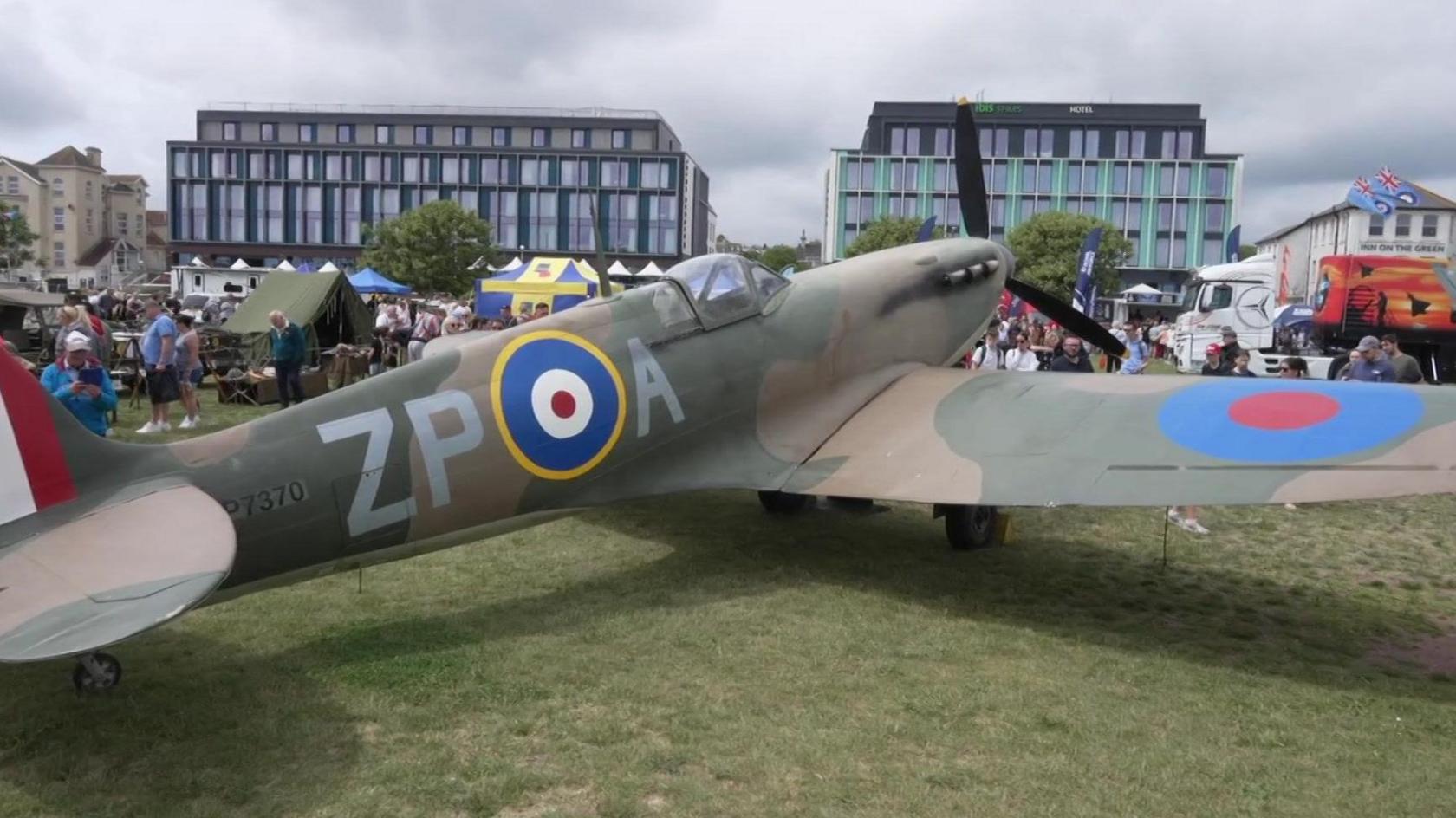 A replica Spitfire plane on display on Paignton Green during the second day of the English Rivera Airshow. People are walking around the site which has a number of display tents set up.